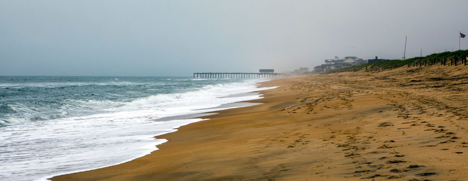 A wide empty stretch of dune-backed Atlantic beach with a fishing pier in the misty distance