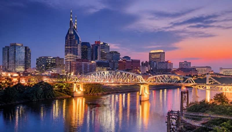 The Nashville skyline and pedestrian bridge lit up at dusk, reflected in the Cumberland River