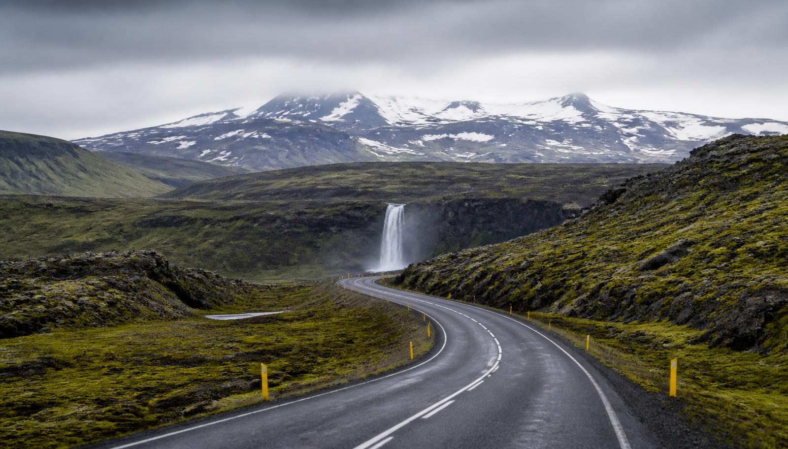 An empty two-lane road curving through green Icelandic hills toward a snow-capped volcano and waterfall